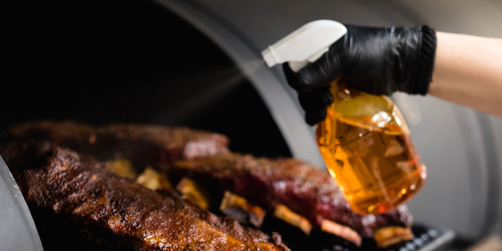 BBQ expert spritzing meat with an apple cider vinegar meat spritz ribs on a grill